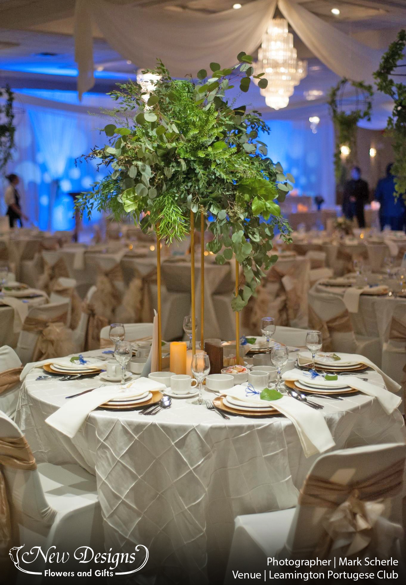 A Centerpiece of wild greenery sits atop a gold 4 posted metal frame. Atop white linen. Chairs covered with fitted white spandex, and a trailing textured cream accent swirling down the back.
